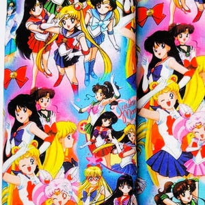 May include: A colorful Sailor Moon-themed wristlet and two matching fabric pouches. The wristlet is a narrow strap with a silver metal clasp. The pouches are rectangular and feature a vibrant print of Sailor Moon characters in various poses.