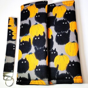 May include: A fabric wristlet keychain and two rectangular fabric covers featuring a repeating pattern of fluffy black cats and bright orange pumpkins on a gray background. The wristlet includes a silver metal clasp and a circular split key ring.