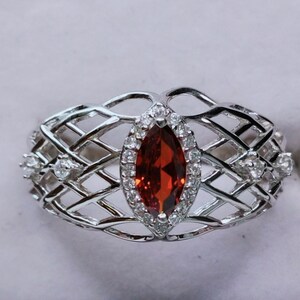 May include: A silver ring featuring a red marquise-shaped gemstone surrounded by smaller clear gemstones. The ring has a delicate, intricate design.