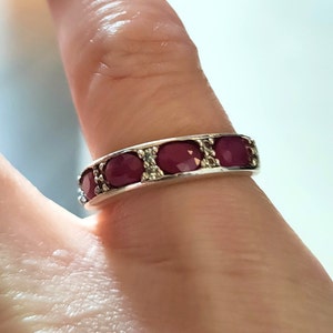 以下が含まれることがあります： 5つの赤い宝石が並んだ銀のバンドリング。リングは指にはめられています。