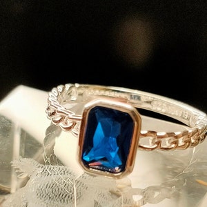 Puede incluir: Un anillo de plata con una piedra preciosa azul engastada en un bisel cuadrado. La banda es un diseño de eslabones de cadena.