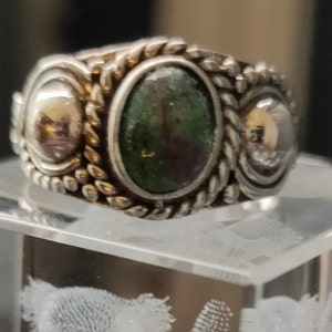 May include: A silver ring with a green gemstone and two smaller silver accents. The ring has a braided design around the edge.