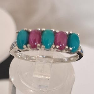 May include: A silver ring with five oval gemstones. The gemstones alternate between turquoise and magenta. The ring is displayed on a clear stand. The ring's band is marked with '925'. The gemstones are set in a row across the top of the ring.