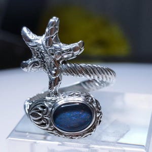 May include: A silver ring featuring a detailed starfish design and a blue opal gemstone. The ring is a statement piece with a unique and intricate design.