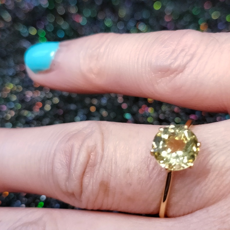 May include: A gold ring with a round, yellow gemstone set in a bezel setting. The ring is on a finger with light blue nail polish.