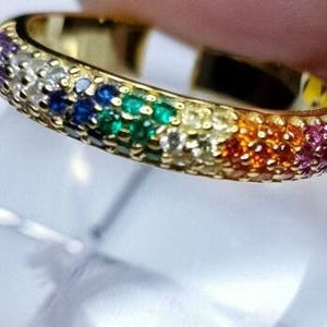 Könnte beinhalten: Ein goldfarbener Ring mit einem Band, das mit kleinen, bunten Edelsteinen bedeckt ist. Die Edelsteine sind in einem Regenbogenmuster angeordnet und zeigen Farben wie Lila, Weiß, Blau, Grün, Gelb, Orange und Rosa. Der Ring wird im Nahaufnahme gezeigt.