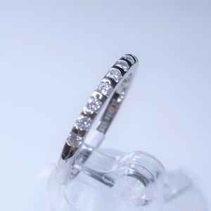 Pode incluir: Uma banda de prata com uma fileira de pequenos diamantes transparentes engastados em uma linha contínua ao redor da banda.