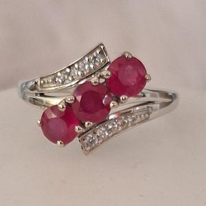 May include: A silver-toned ring featuring three round, deep red rubies set in a diagonal row. The band is accented with two curved channels containing small, clear, sparkling stones that flank the central rubies on both sides of the setting.