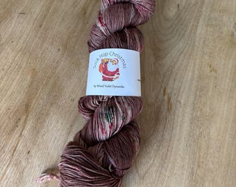 May include: A skein of Sock Hop Christmas yarn by Wood Violet Dyeworks. The yarn is a mix of burgundy, cream, and red, with a label featuring a Santa Claus illustration. The yarn is wound into a compact bundle, ready for knitting or crocheting.