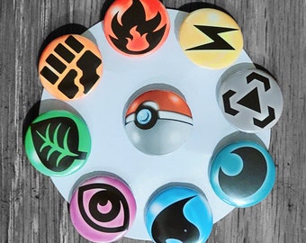 Può includere: Nove bottoni rotondi con diversi simboli di tipo Pokémon. I simboli sono fuoco, elettrico, lotta, normale, erba, psichico, fantasma, acqua e drago. I bottoni sono disposti in cerchio su uno sfondo bianco.