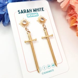 May include: Gold-colored sword earrings with a sunburst design at the top, hanging from a small chain. The earrings are displayed on a white card with the text "SARAH WHITE DESIGNS" and social media icons.