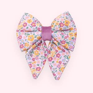 May include: A large, fabric hair bow with a floral pattern in yellow, pink, and blue on a white background. The center of the bow is a solid, light purple velvet. The bow is a decorative hair accessory.