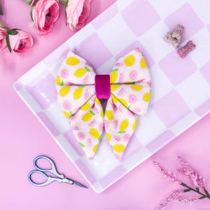 Pode incluir: Uma gravata borboleta decorativa com um padrão de limões amarelos e rosa. A gravata borboleta tem um centro magenta e está colocada numa placa xadrez rosa e branca. Flores rosas e uma tesoura também estão presentes.