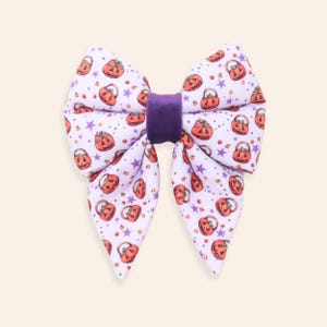 Può includere: Un fiocco per capelli in tessuto bianco con un motivo ripetuto di secchielli per caramelle a forma di zucca arancione e piccole stelle viola. Il centro del fiocco è stretto da una fascia in velluto viola scuro, creando una forma strutturata.