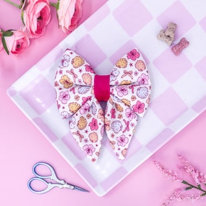 May include: A large, fabric bow with a seashell and floral pattern in pink, yellow, and white, centered with a bright pink band. The bow sits on a pink and white checkered plate, with pink flowers and scissors nearby.