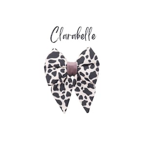 May include: A large, black and white cow print bow with a mauve velvet center. The bow is tied in a classic bow shape and is ready to be worn in hair.