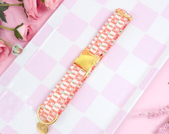 May include: A pet collar featuring a repeating pattern of white bunnies and orange carrots on a light pink and white checkered background. The collar includes a gold-toned metal side-release buckle and a gold-toned D-ring with a small circular tag.