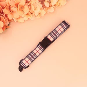 May include: A dog collar with a beige, red, and navy blue plaid design. It has a black plastic buckle and a metal loop for attaching a lead or tag. The collar is set against a peach background.