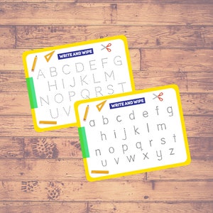 May include: Two write-and-wipe educational boards with the alphabet in uppercase and lowercase letters. The boards have yellow borders, green accents, and include a pencil and scissors graphic. The background is a wooden surface.