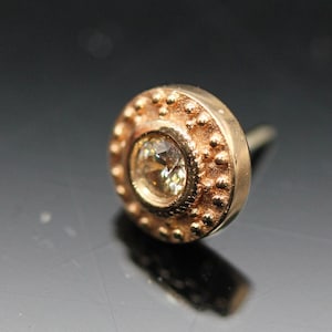 May include: A gold stud earring with a round, clear gemstone set in a textured bezel. The earring has a post and a small, round back.