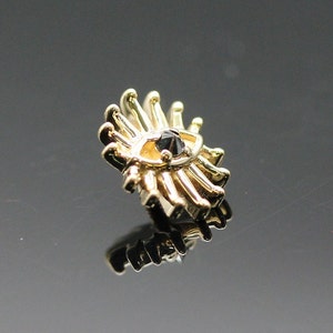 May include: Gold body jewelry with a black gemstone in the center, shaped like an eye with radiating lines.