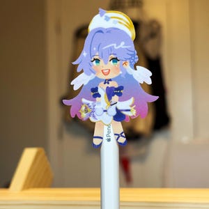 K&ouml;nnte beinhalten: Ein farbenfroher Aufkleber im Anime-Stil mit einem wei&szlig;en und gelben Heiligenschein, lila Haaren und blauen Augen. Der Aufkleber ist an einem wei&szlig;en Bleistift befestigt. Die Figur tr&auml;gt ein wei&szlig;es Kleid mit blauen Akzenten und hat kleine Fl&uuml;gel.