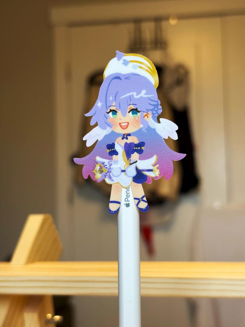 K&ouml;nnte beinhalten: Ein farbenfroher Aufkleber im Anime-Stil mit einem wei&szlig;en und gelben Heiligenschein, lila Haaren und blauen Augen. Der Aufkleber ist an einem wei&szlig;en Bleistift befestigt. Die Figur tr&auml;gt ein wei&szlig;es Kleid mit blauen Akzenten und hat kleine Fl&uuml;gel.