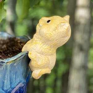 May include: A yellow ceramic animal figurine shaped like a cat, with a black eye, is perched on the edge of a blue and white ceramic planter.