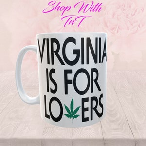 Może przedstawiać: Biały ceramiczny kubek z czarnym napisem "VIRGINIA IS FOR LOVERS", z zielonym liściem konopi zastępującym "O" w słowie "LOVERS". Kubek ma zakrzywiony uchwyt, a nad napisem różowe słowa "Shop With TnT".