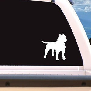 May include: A white vinyl decal of a dog silhouette is affixed to the rear window of a white pickup truck. The dog is a muscular breed, standing in profile. The background shows a sky with clouds.