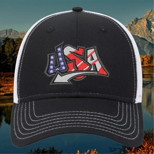 Puede incluir: Una gorra de camionero negra y blanca con un gráfico bordado "USA" en rojo, blanco y azul. La gorra tiene una parte trasera de malla y una visera curvada.