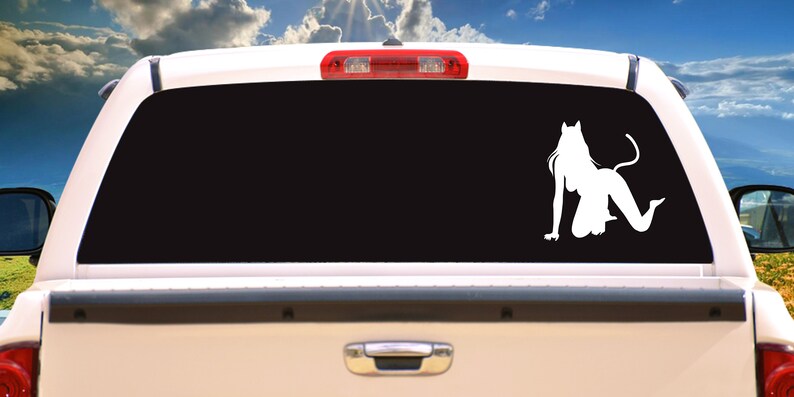 K&ouml;nnte beinhalten: Wei&szlig;es Vinyl-Aufkleber einer knienden Figur mit Katzenohren und Schwanz, angebracht an der Heckscheibe eines wei&szlig;en Pick-ups. Die Figur ist eine Silhouette vor der schwarzen T&ouml;nung, mit einem Hintergrund aus blauem Himmel und gr&uuml;nen H&uuml;geln.