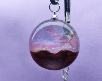 May include: A round, clear resin pendant necklace with a pink and purple galaxy-like design and a dark brown and gold mountain range at the bottom.