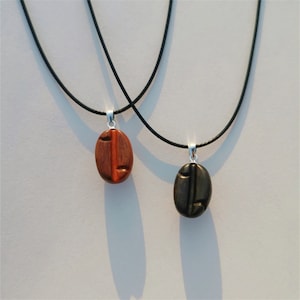 Può includere: Due collane con cordoncini neri e ciondoli in legno. I ciondoli sono scolpiti a forma di chicco di caffè. Un ciondolo è marrone e l'altro è nero.