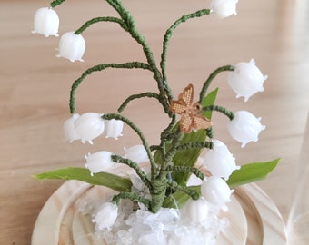 May include: A glass dome with a white lily of the valley flower arrangement inside. The flowers are made of white beads and the stems are green wire. There are two gold butterflies on the stems. The base of the dome is a wooden circle.