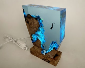 Könnte beinhalten: Eine Harzlampe mit blauem und grünem Leuchten, die eine Silhouette eines Tauchers im klaren Harz zeigt. Die Lampe ist auf einem Holzsockel montiert.