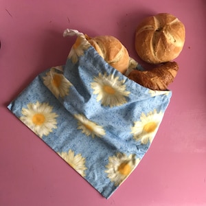 Op de afbeelding: Een blauwe herbruikbare boodschappentas met een bloemenpatroon met gele madeliefjes. De tas is gevuld met drie gebakken producten: een croissant, een rond broodje en een brood.