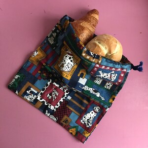 Op de afbeelding: Een blauwe en bruine herbruikbare boodschappentas met een dalmatiër-hondenprint. De tas heeft een trekkoord sluiting en is gevuld met twee broden.