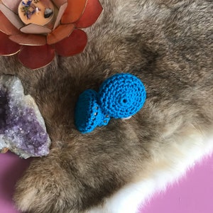 May include: Two small crocheted blue mushroom-shaped ornaments on a brown faux fur background.
