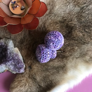May include: Two purple crocheted balls with a silver pin attached. The balls are on a brown and white fur surface.