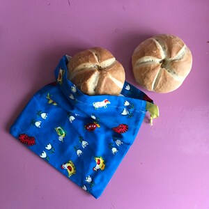 Op de afbeelding: Twee ronde broodjes, een in een blauwe stoffen tas met een wit en geel bloemenpatroon. De tas heeft een gele trekkoord.