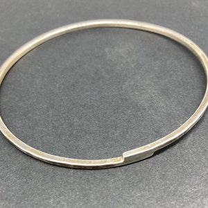 May include: A silver bangle bracelet with a simple, open design. The bracelet is made of a thin, flat band of metal that is slightly curved. The ends of the bracelet are joined together with a small, flat piece of metal.