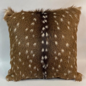 May include: A square decorative pillow with a brown and white spotted pattern, resembling deer fur. The pillow has a central dark brown stripe and a fluffy, textured border. The pillow is a home decor item.