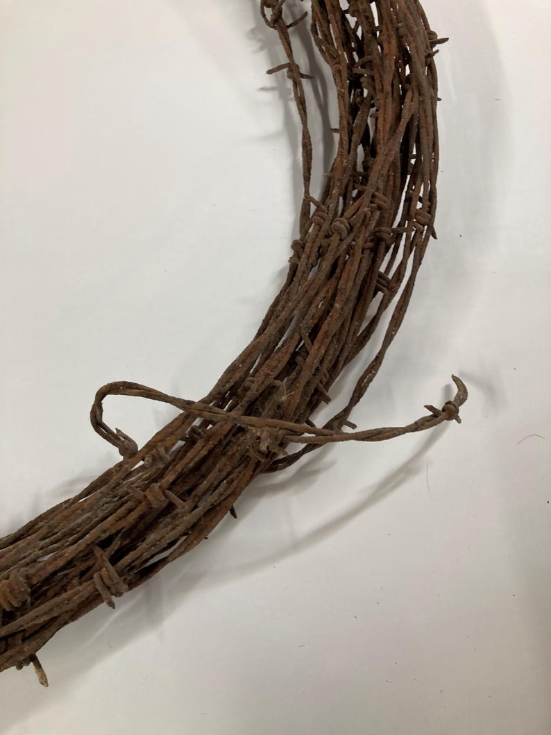 May include: Close-up of a rustic, circular wreath made of weathered, rusty barbed wire. The brown metal wire is tightly wound, with sharp barbs protruding. The wreath is set against a plain white background, highlighting its texture and color.