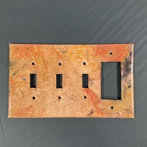 May include: A copper-colored light switch plate with a marbled pattern in shades of orange, red, and brown. The plate has three toggle switches and one rectangular outlet.