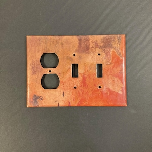 May include: A copper-colored outlet and light switch cover plate with a rustic, distressed finish. The plate has a mottled orange and brown color with a hint of red.