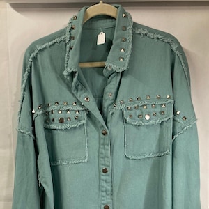 May include: A light teal denim jacket with silver-colored stud embellishments. The jacket features a classic collar, button-down front, two chest pockets, and frayed edges. The jacket is hanging on a wooden hanger.