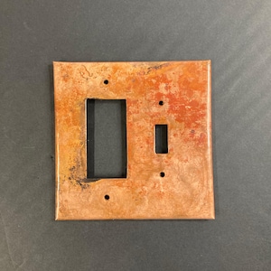 May include: A copper-colored light switch and outlet cover plate with a rustic, distressed finish. The plate has two openings, one for a standard light switch and one for an outlet.
