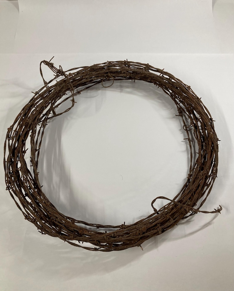 May include: A circular wreath made of rusty brown barbed wire. The wire is tightly wound, creating a dense, textured appearance. The wreath is set against a plain white background, highlighting its rustic aesthetic.