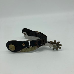 May include: A pair of silver spur straps with black leather and a star-shaped spur.